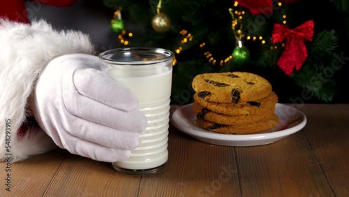 Santa Claus eating cookie and milk. Santa hands picking cookie and milk glass on wooden table against festive Christmas tree,