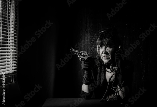 Monochrome shot of an elderly woman in a black veil talking on the phone with a gun in her hand