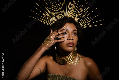 tattooed african american woman with golden crown and paint on face looking up isolated on black.