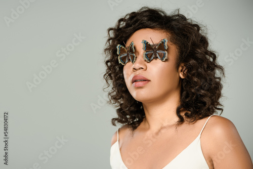 curly african american woman with butterflies on eyes isolated on grey.