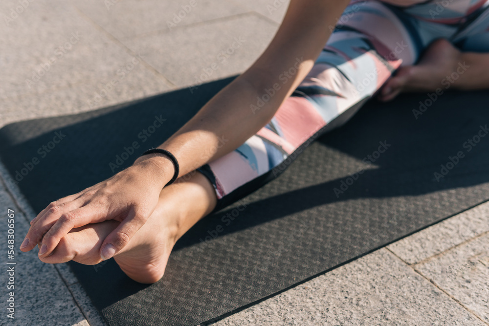 Cropped shot of a woman doing leg stretches grabbing her foot with her leg outstretched and resting on the yoga mat in the street.