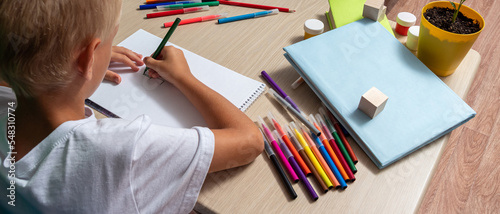 Boy draws with felt-tip pen on paper. On table there are pencils, brush, book, cubes, paints. Free time, learning to draw, hobbies. Close-up. Selective focus.