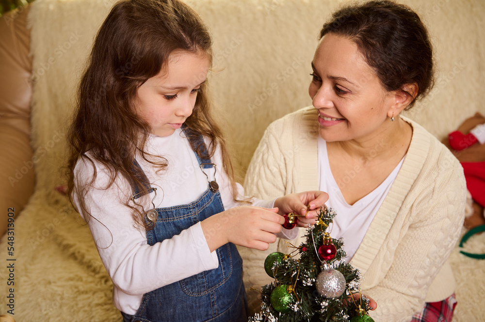 Overhead view of a loving affectionate mother hugging her adorable little daughter, who is hanging shiny toy balls and bauble on a small Christmas tree, making preparations for celebrating New Year
