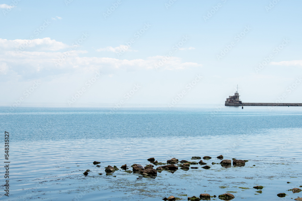 View of seaport in city against cloudy sky. Port cranes are located along the coastline. In foreground is emerald water of Black Sea. Feodosia port