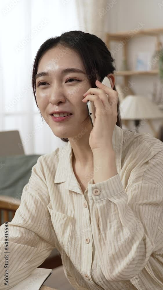 Vertical Screen: half length shot of a happy asian japanese business woman chatting on the cell phone with a smiling face while working from home in a bright living room. Vertical Screen: half length shot of a happy asian japanese business woman chatting on the cell phone with a smiling face while working from home in a bright living room.