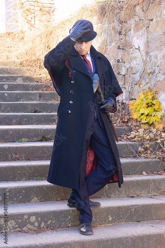 A young respectable man in a vintage coat, suit, bowler hat and with a cane is standing on the stairs