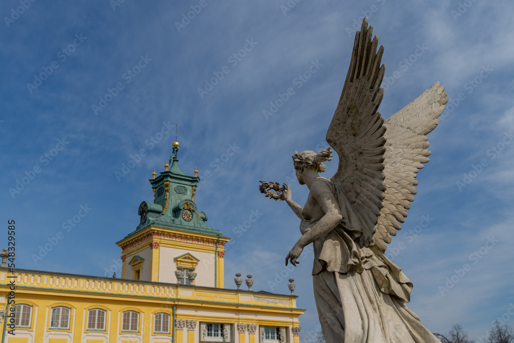 the statue of an angel holds a wreath in his hand directed towards the Wilanowski Palace in Warsaw, the statue is against the background of the blue sky, a view from below