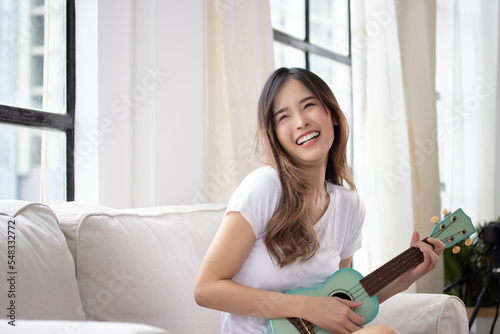 Happy asian women enjoy singing by playing guitar, ukulele while sitting on sofa at home.