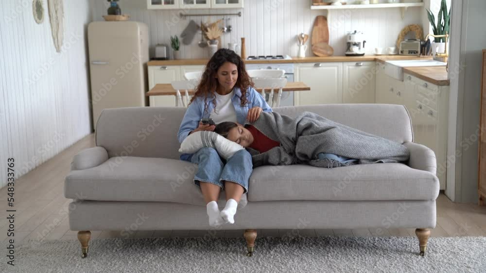 Loving Caucasian mother and teen girl sleeping on lap located on sofa in their apartment. Long-haired woman spending time with daughter simultaneously using mobile phone for shopping in online store 