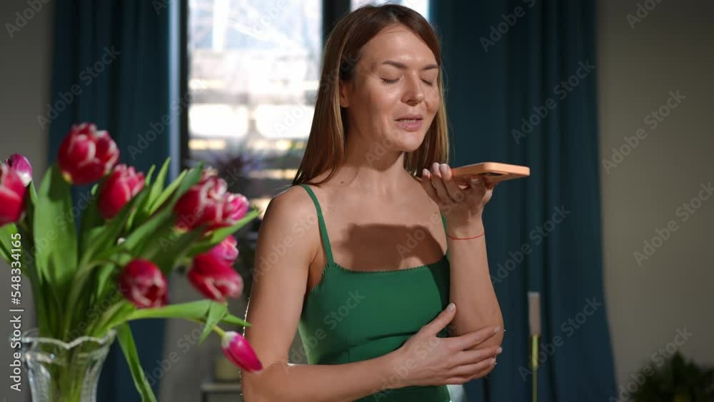 Brunette young woman in green dress recording voice message on smartphone thanking for flowers. Portrait of happy satisfied Caucasian slim beautiful lady talking smiling standing indoors with tulips Brunette young woman in green dress recording voice message on smartphone thanking for flowers. Portrait of happy satisfied Caucasian slim beautiful lady talking smiling standing indoors with tulips