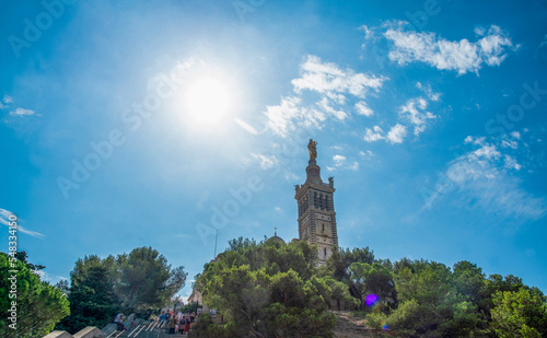 Amazing view on Basilique Notre-Dame de la Garde in Marseille and tourists blurred motion and awe pine trees and sun shining in blue sky