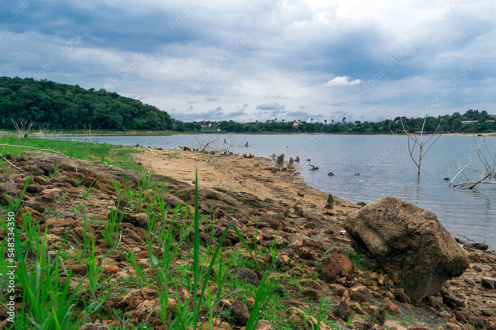 Linda vista da orla da lagoa Várzea das Flores em contagem, com muitas pedras e vegetação rasteira, céu com muitas nuvens e construções e matas ao fundo. Linda vista da orla da lagoa Várzea das Flores em contagem, com muitas pedras e vegetação rasteira, céu com muitas nuvens e construções e matas ao fundo.