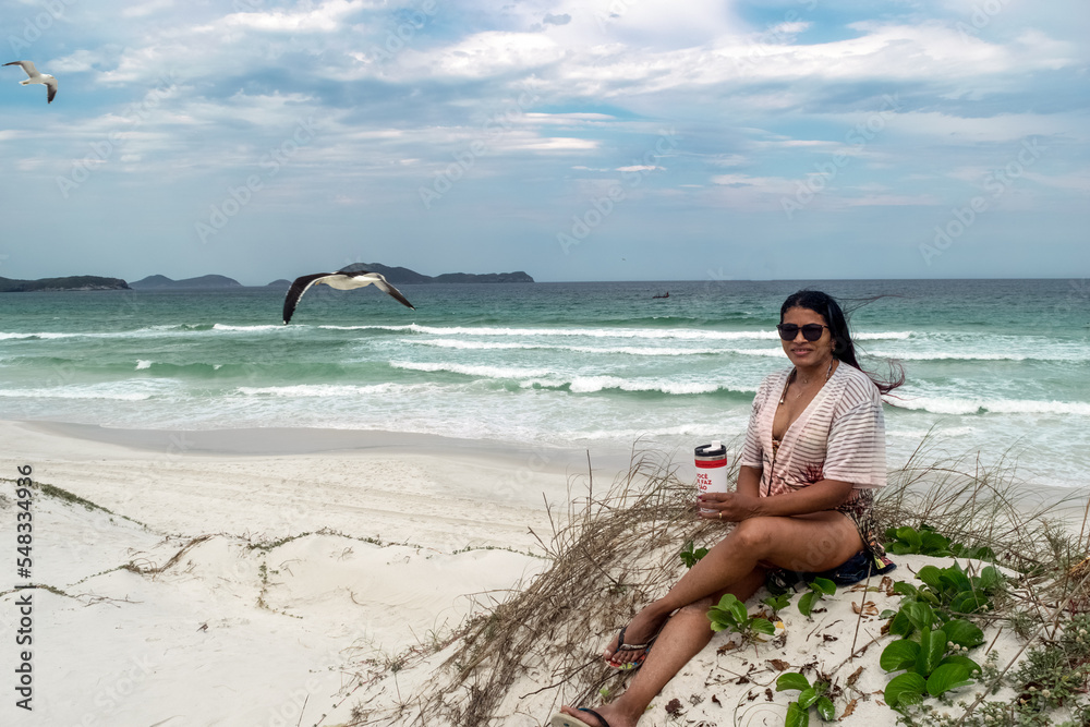 Mulher na Orla da Praia das Dunas, sentada em uma das dunas de areia, com pássaros em volta e céu azul com nuvens.