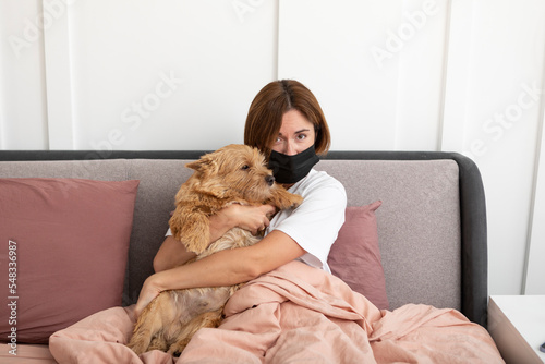 Stressing woman wearing medical mask sits on the bed with her cute pet dog. Allergy and illness concept