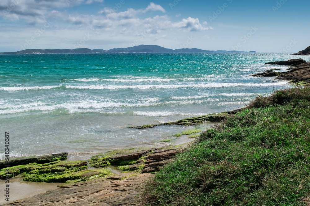 Vista da linda praia das Conchas, próxima a cidade de Cabo Frio, com mar azul em volta, vegetação rasteira, rochas com musgo e montanhas ao fundo.