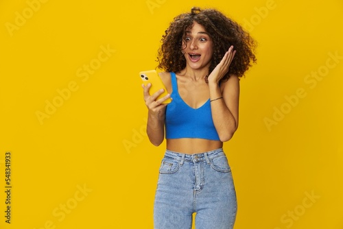 Influencer woman with phone in hand taking selfies talking on video call freelancer social media technology work, with curly afro hair in blue shirt yellow background , smile with teeth, copy space