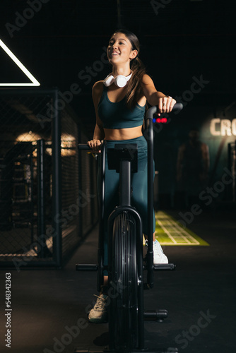 An Asian woman in a gym exercising on a bicycle. Women making an effort while exercising on an air bike. The girl is wearing headphones.