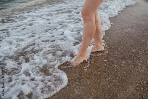 Slim female legs and feet walking along sea water waves on sandy beach. Pretty woman walks at seaside surf. Splashes of water and foam. Girl after bathing in ocean go on shore.