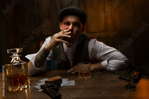 Young gangster smoking at table with cards and gun in vintage casino