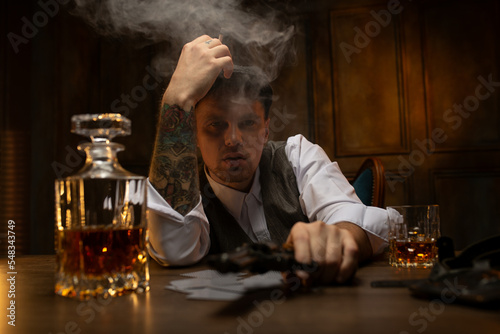 Young gangster smoking and holding revolver at table with cards and decanter of whiskey photo