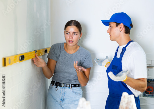 Bild auf Leinwand Girl dissatisfied with quality of renovation works carried out in her apartment, checking flatness of wall surface with spirit level and talking to confused young builder
