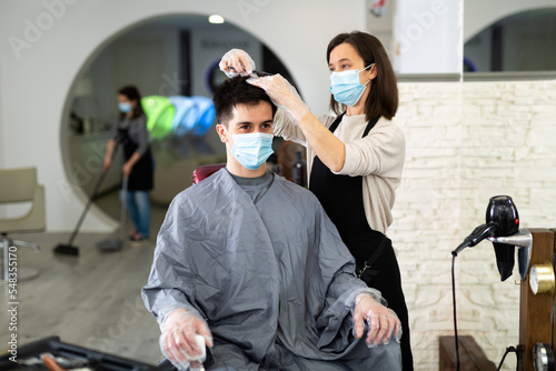 Female hairdresser making hair styling for young man in protective mask and gloves, working day during pandemic situation