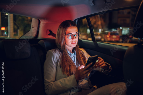 Tablou pe pânză Business woman using smartphone while sitting in a backseat of a car at night