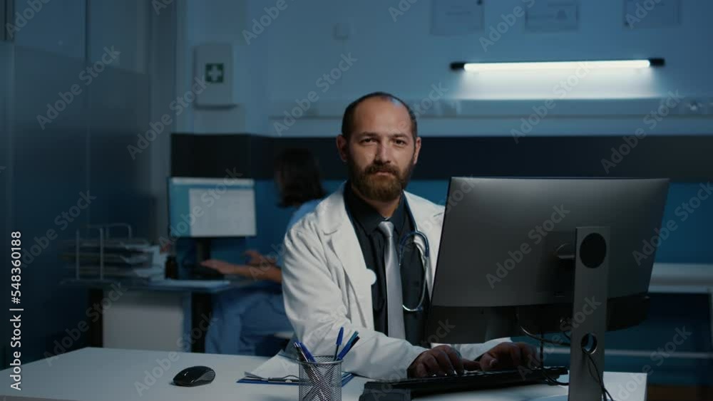 Specialist doctor in medical coat smiling at camera while analyzing patient expertise typing health care treatment on computer working after hours in hospital office. Medicine concept Specialist doctor in medical coat smiling at camera while analyzing patient expertise typing health care treatment on computer working after hours in hospital office. Medicine concept