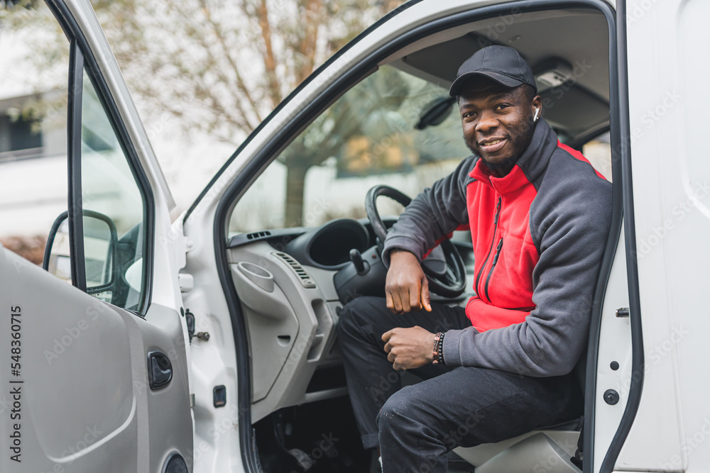 Full of energy, positive African-American deliveryman posing on driver's seat with open door. Courier outfit - black hat and red-and-gray jacket. Outdoor shot. High quality photo Full of energy, positive African-American deliveryman posing on driver's seat with open door. Courier outfit - black hat and red-and-gray jacket. Outdoor shot. High quality photo