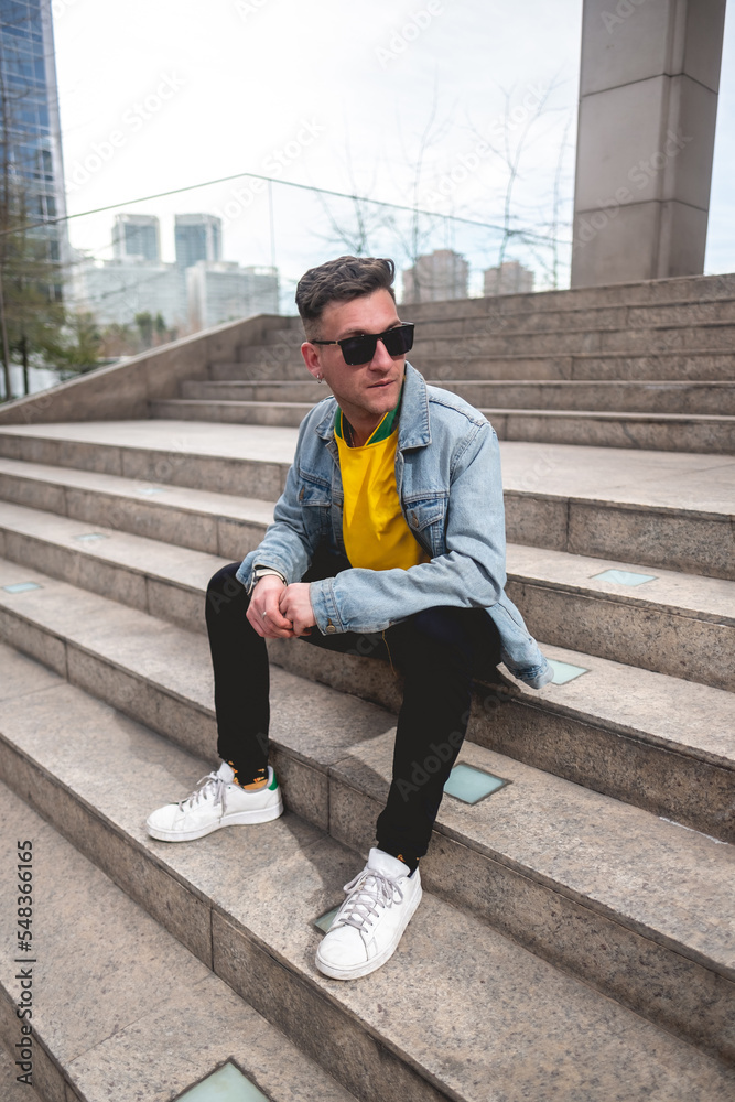 Young blonde model guy with sunglasses, yellow t-shirt, denim jacket and black jeans sitting on stone stairs in a park in the city in the afternoon Young blonde model guy with sunglasses, yellow t-shirt, denim jacket and black jeans sitting on stone stairs in a park in the city in the afternoon