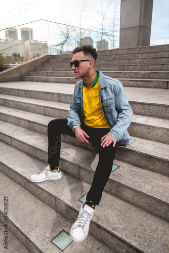 Young blonde model guy with 4-leaf clover tattoo and sunglasses  yellow t-shirt  denim jacket and black jeans sitting on stone stairs in a park in the city in the afternoon