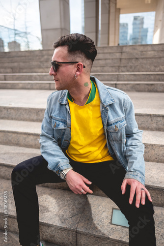 Young blonde model guy with 4-leaf clover tattoo and sunglasses, yellow t-shirt, denim jacket and black jeans sitting on stone stairs in a park in the city in the afternoon