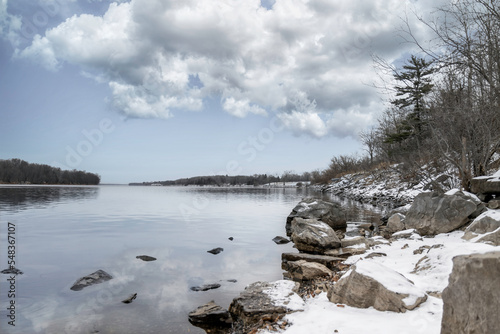 Calm river on a winter morning, rocky shoreline with snow, trees on shore, clouds reflected in water, nobody