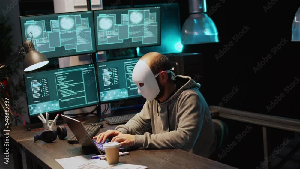 Cyber impostor wearing mask hacking database servers, hacker with hood breaking into computer system and activating virus to create malware. Mysterious impostor stealing big data. Handheld shot.