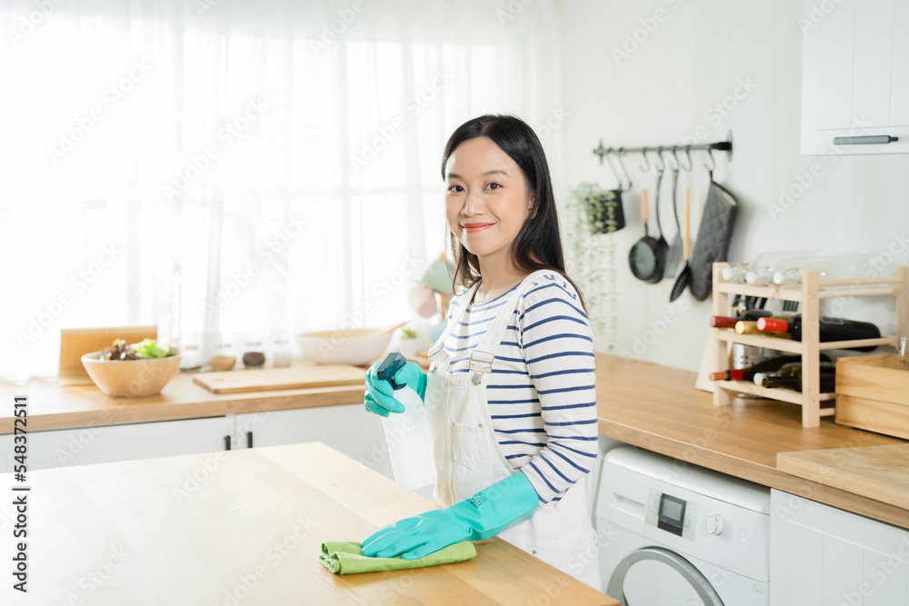 Young Asian woman looking at camera while spraying and cleaning the table with rag cloth. Beautiful cleaning service worker housekeeping and tidying up the kitchen. Housework and chores concept..