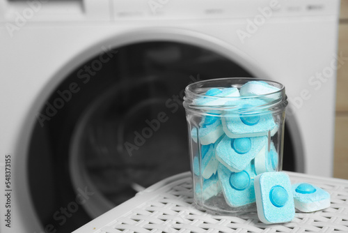 Jar with water softener tablets on laundry basket near washing machine, space for text photo