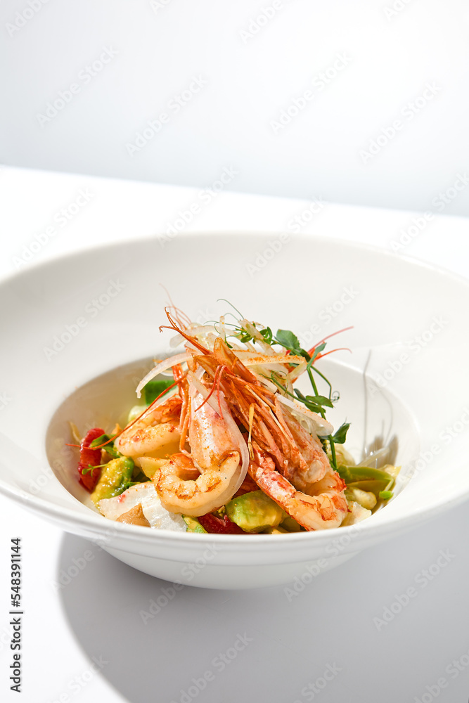 Elegant food - salad with shrimp and avocado on white table with harsh shadow. Flashy food concept. Seafood salad with shrimp, tomatoes and avocado on white plate with sunlight. Elegant food - salad with shrimp and avocado on white table with harsh shadow. Flashy food concept. Seafood salad with shrimp, tomatoes and avocado on white plate with sunlight.