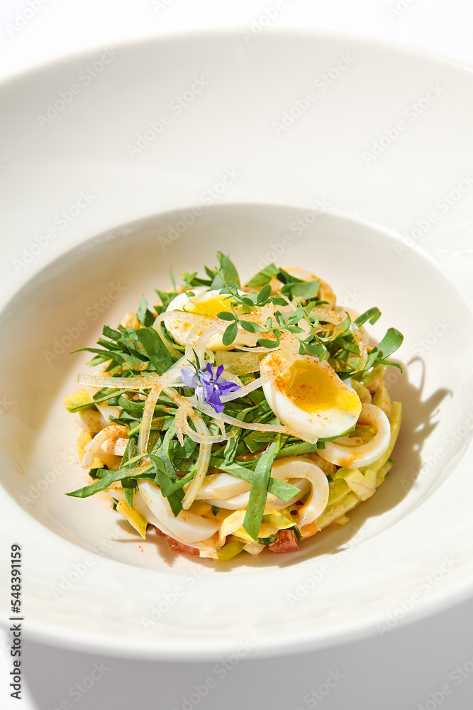 Healthy salad with squid and avocado on white plate with harsh shadows. Calamari salad with vegetables and eggs on white background with shadows of leaves. Summer food menu.