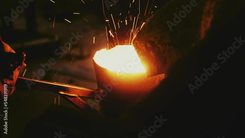 Professional blacksmith works with red-hot melted iron from cast-iron furnace at industrial steel factory. Unrecognizable man uses fireproof tools closeup