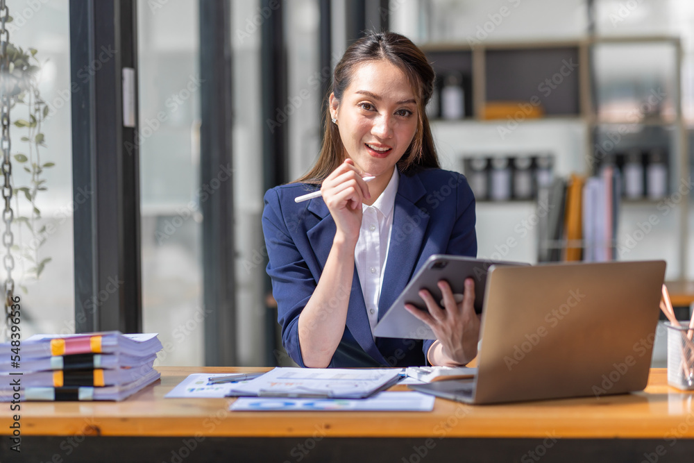 Brown haired wearing light blue jacket smiling Asian woman work with document laptop in office, doing planning analyzing the financial report, business plan investment, finance analysis concept. Brown haired wearing light blue jacket smiling Asian woman work with document laptop in office, doing planning analyzing the financial report, business plan investment, finance analysis concept.