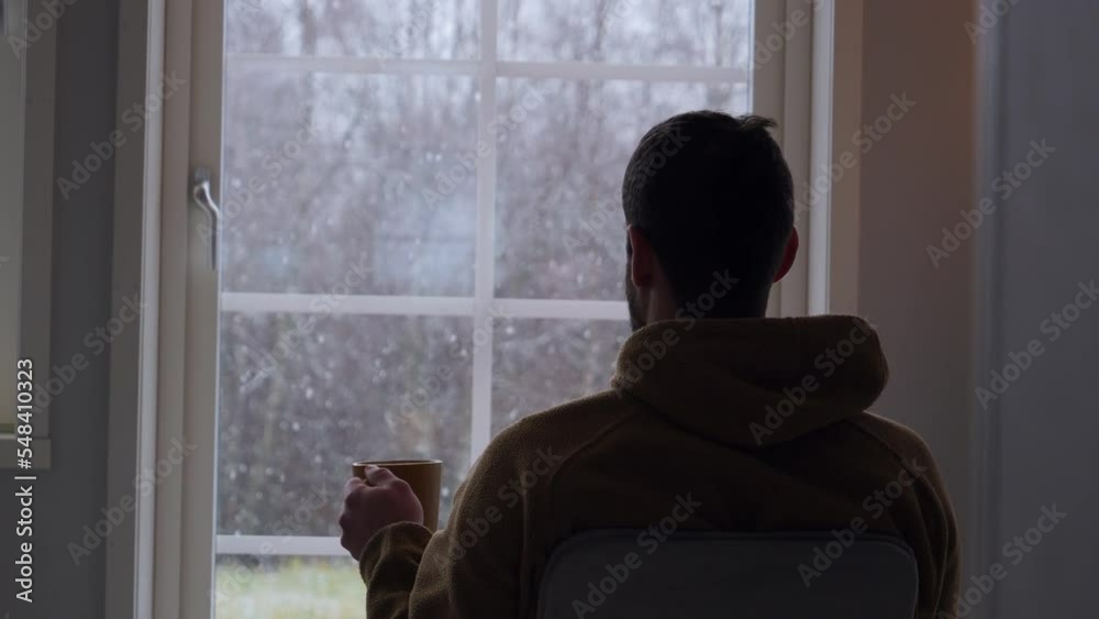 Man drinking tea and seeing first autumn snow through house window. Zoom in Man drinking tea and seeing first autumn snow through house window. Zoom in