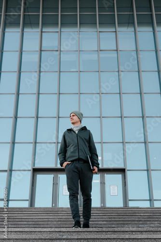 Young man in outerwear holding a laptop standing on the steps against the backdrop of a modern building and looking away