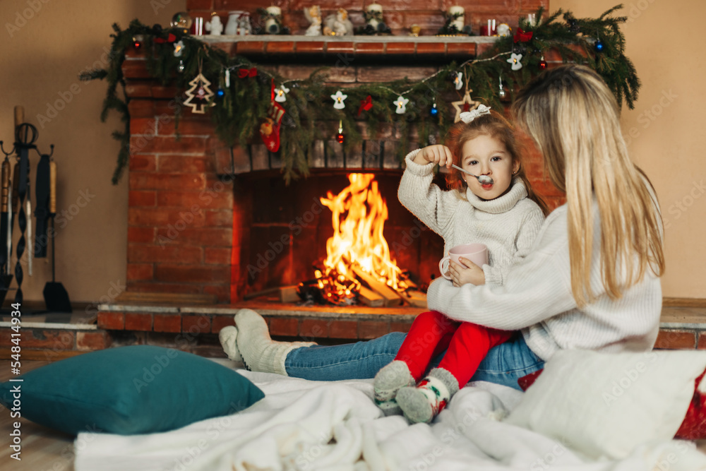 Family mother and child daughter hug and warm up on a winter evening by the fireplace, drink hot cocoa. Christmas Eve or New Year's Eve, Christmas holidays, Christmas time, family evening