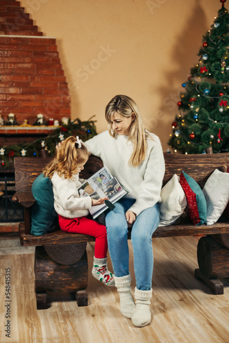 Cute mom and daughter are looking through a family photo album on Christmas Eve, spending time together by the fireplace and the Christmas tree. Christmas, New Year, memories, family evening