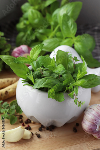 Mortar with different fresh herbs near garlic, horseradish roots and black peppercorns on wooden table, closeup