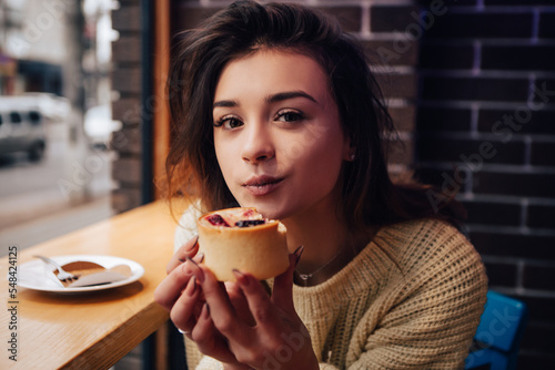 Smiling woman dressed warm sweater  enjoy eating dessert  tasty cupcake. Snack food in street cafe. New Year Christmas decorations  garlands 