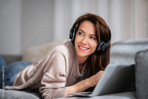 Attractive and cheerful woman using laptop and headphone while relaxing at home