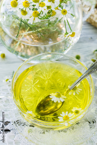 Chamomile herbal tea in a glass cup on white crochet napkin on wooden table, healthy chamomile flower drink with honey, fresh chamomile flowers in background, healthcare and healthy eating concept