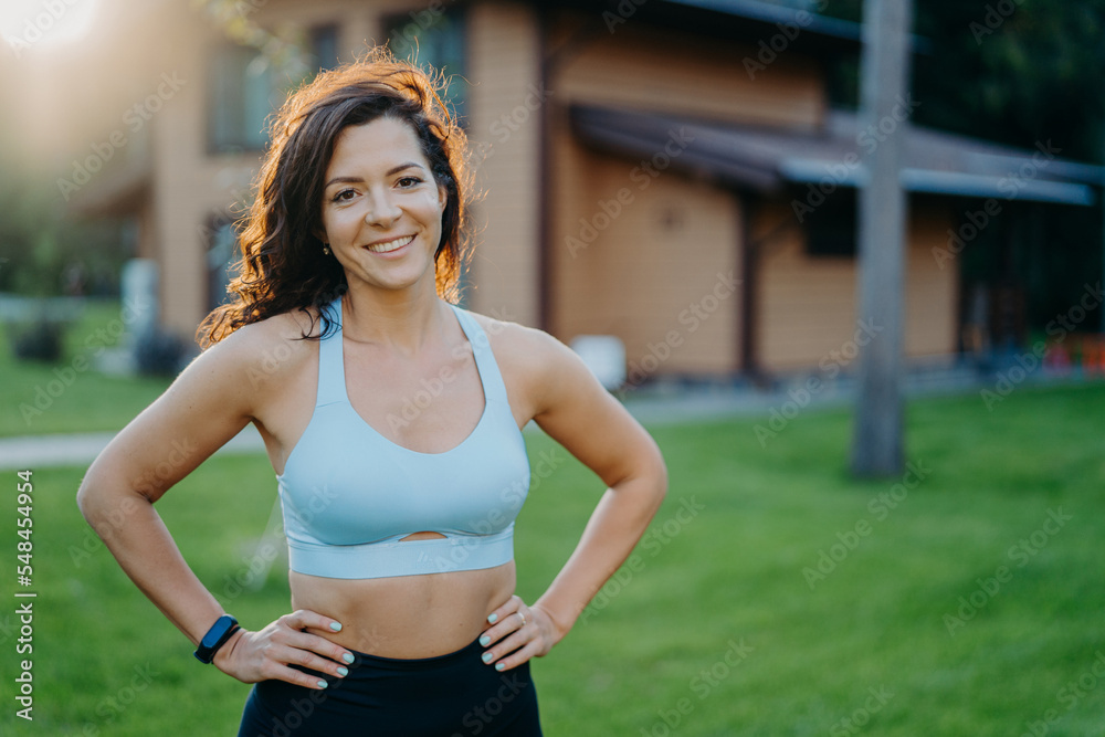 Beautiful pleased curly haired woman in good shape, keeps hands on waist, dressed in cropped top, smiles gladfully, has sporty body, poses outdoor against house background. Morning workout concept Beautiful pleased curly haired woman in good shape, keeps hands on waist, dressed in cropped top, smiles gladfully, has sporty body, poses outdoor against house background. Morning workout concept
