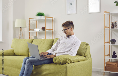 Serious young man in glasses sitting with his notebook PC on couch at home. Remote student or corporate employee working on modern laptop computer while resting on comfortable sofa in living room